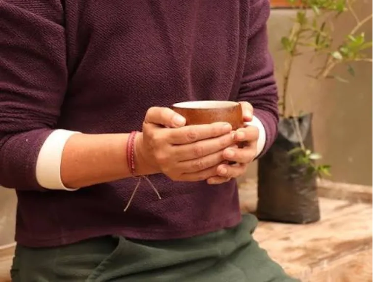 Close-up of two hands holding a clay cup, with a person in a maroon sweater in the background.